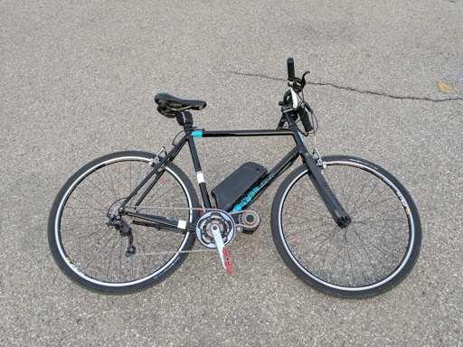 Black electric bicycle on gray asphalt pavement, featuring a battery pack mounted on the frame, front and rear wheels with silver rims, black handlebars with brake levers, a black saddle, and visible gear mechanism.