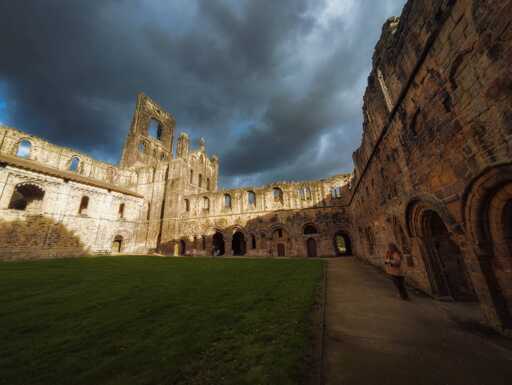 A dramatic view of the ruins of Kirkstall Abbey taken from the grassy cloister courtyard, looking towards the south side of the nave and the partially collapsed central tower. The ancient stone structure is bathed in a bright patch of sunlight, highlighting its warm orange-brown colours and weathered texture, while the sky above is dominated by heavy, dark, brooding storm clouds. The massive walls, punctuated by rows of arched windows and doorways, frame the scene, with a paved walkway leading around the perimeter. A person wearing a light coat is visible in the lower right corner, walking along the wall, providing a sense of scale.