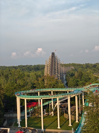 A rollercoaster car cresting the lift hill with the tracks nearly squarely framed in the photo. A log flume is in the foreground.