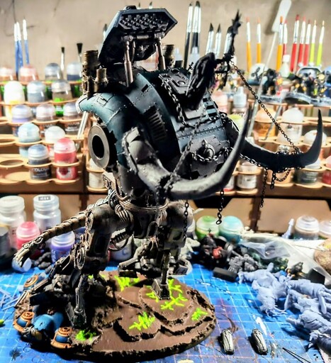 Chaos Acastus Knight Porphyrion model from Games Workshop model posed such that it is recklessly charging forward. The model has been customized to add a mechanical tail and a set of bone tusks protrudes from its carapace armor, as well as a Chaos Sorceress reining in the knight via chains.