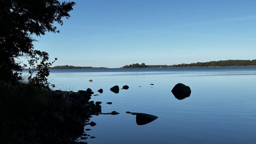 Photo from hiking along a dark shore with some stones sticking up and reflecting black in the calm water. With another forested shore and blue skies on the horizon.