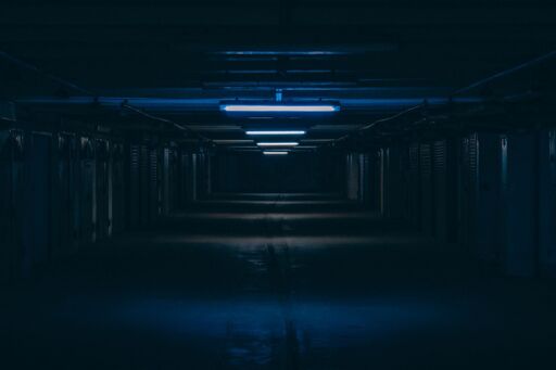 A long, dim corridor lined with identical doors, lit by blue fluorescent lights receding into darkness.
